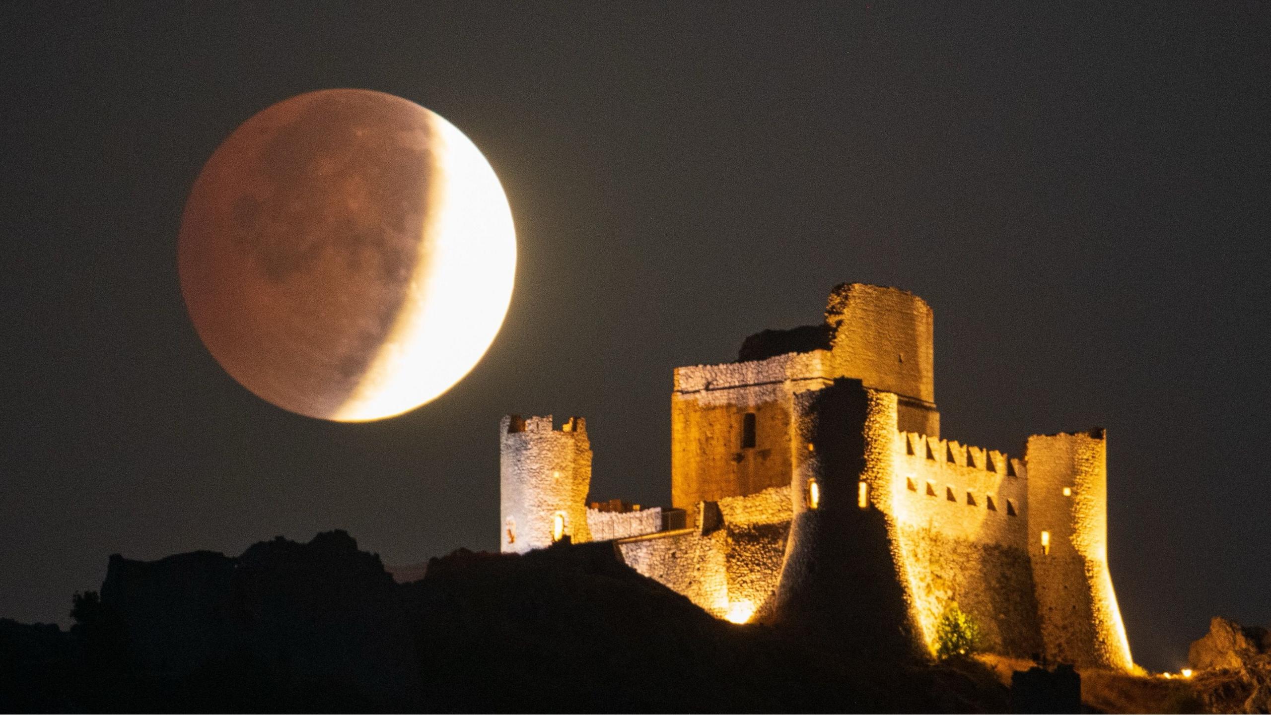A partial lunar eclipse setting over an illuminated fortress