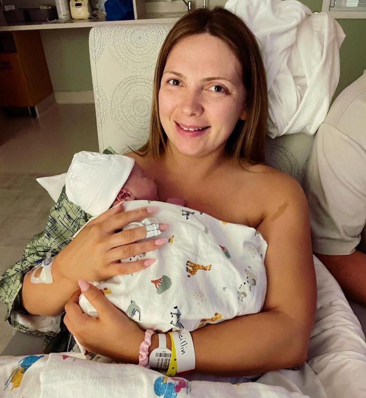 Newborn baby wrapped in a white patterned hospital blanket and wearing a white knit hat, held against the chest of a person seated in a hospital bed. The person has a hospital wristband and an adhesive dressing on one wrist. White bedding and pillows surround the bed. A partially visible person in a white shirt sits beside them. In the background, a hospital room contains a sink, countertop and storage area with medical and personal items.