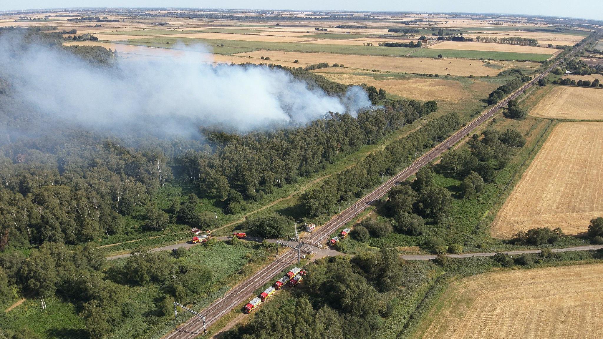 An aerial view of the Holme Fen National Nature Reserve, next to a railway line. Smoke is billowing from the reserve.
