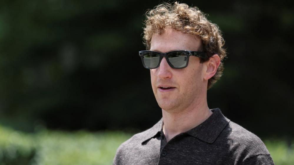 Zuckerberg, wearing black sunglasses and a dark polo shirt, outisde with a green background