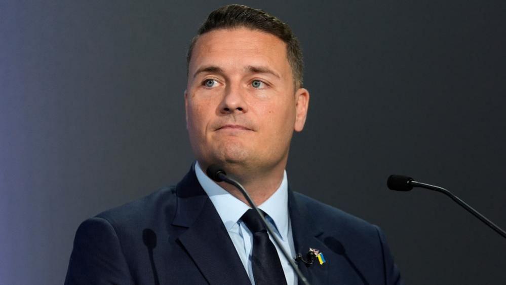 Wes Streeting is stood in front of a microphone. He is wearing a dark-coloured jacket, light blue shirt and a dark-coloured tie. He has short dark hair.