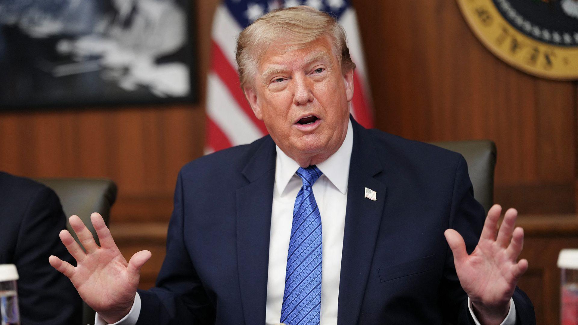 Trump at Camp David in blue jacket and blue tie holding both hands up, pictured in July