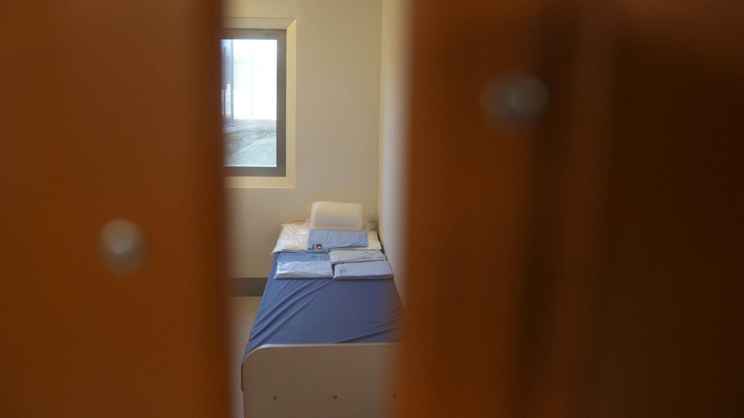 A picture taken from outside a cell looking inside. It is taken through two doors that are not fully opened, creating a glimpse inside. A bed with blue mattress cover in a white room next to a window is showing.