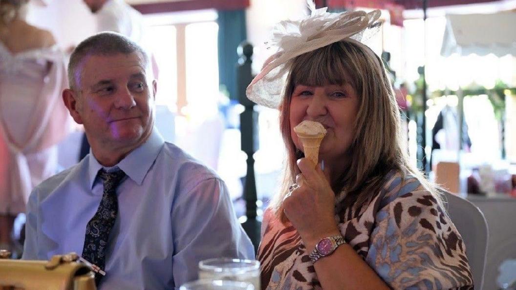 A man sits in a blue shirt and blue tie, to his left (right of picture) a woman sits with a brown and blue dress on and fascinator. She is eating an ice cream in a cone. The background is blurred, but other wedding guests can be seen mingling around.