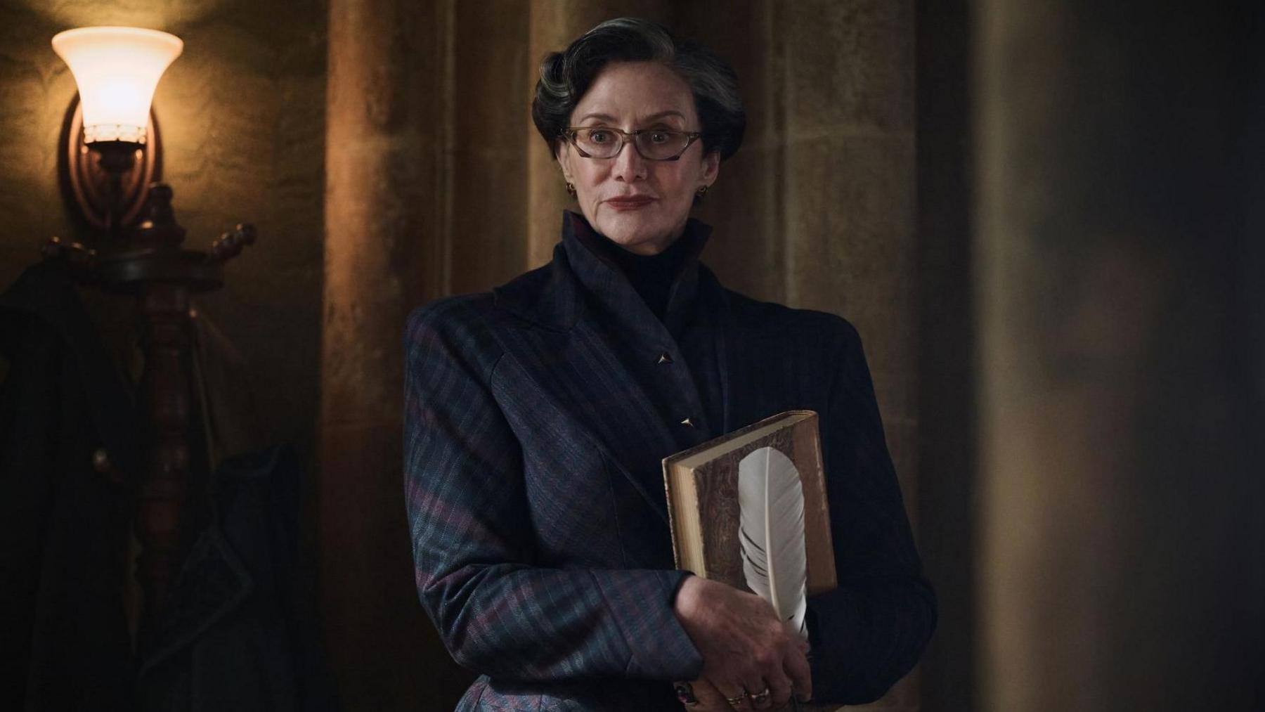 Janet McTeer in a checked jacket holding a book and white feather