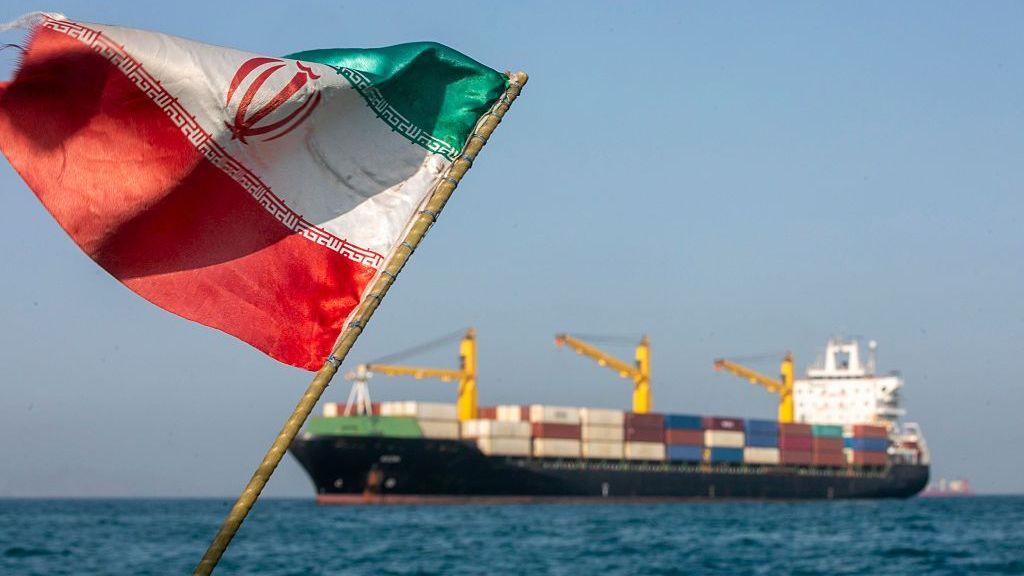 An tattered Iranian flag billowing against a blue sky with the Strait of Hormuz and a large trading ship in the background in soft focus