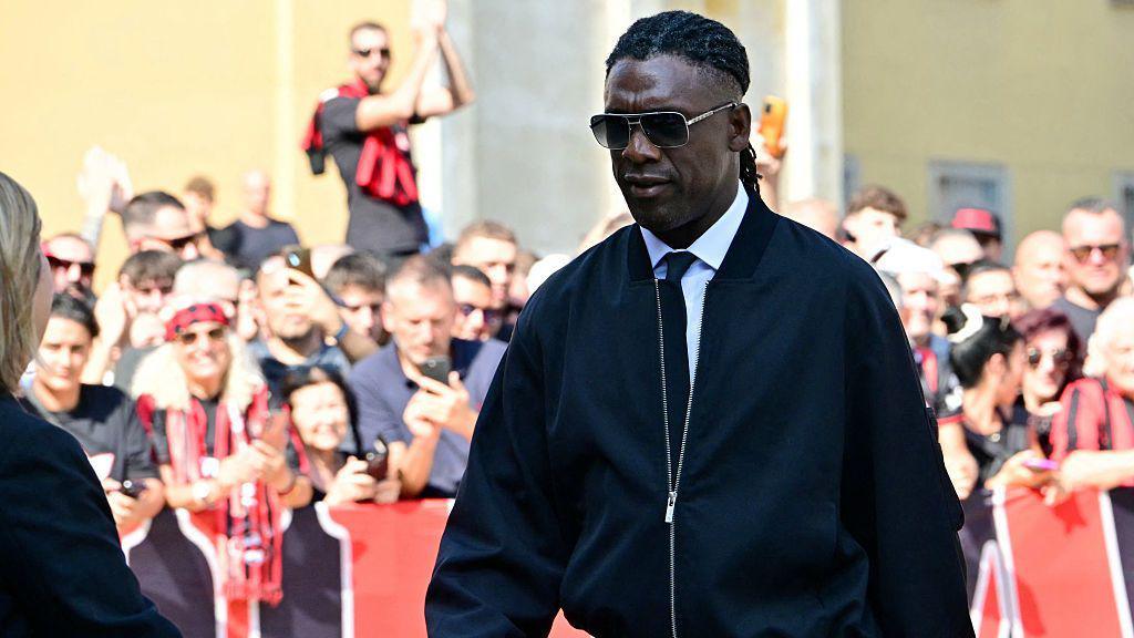 Clarence Seedorf, wearing sunglasses and a black jacket, walks with his head bowed