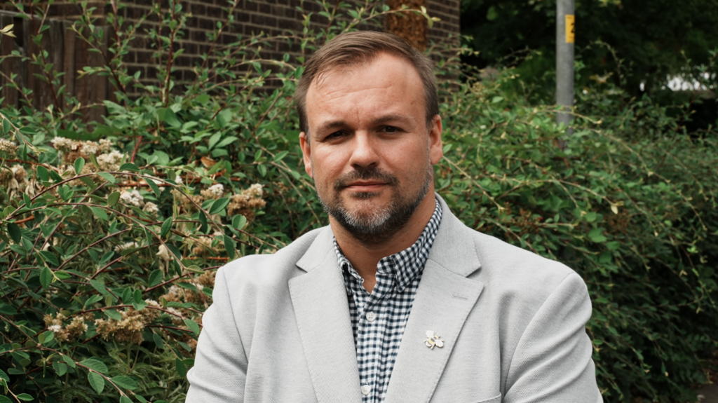 man in grey jacket and checked shirt - he has a beard and wears a lapel pin
