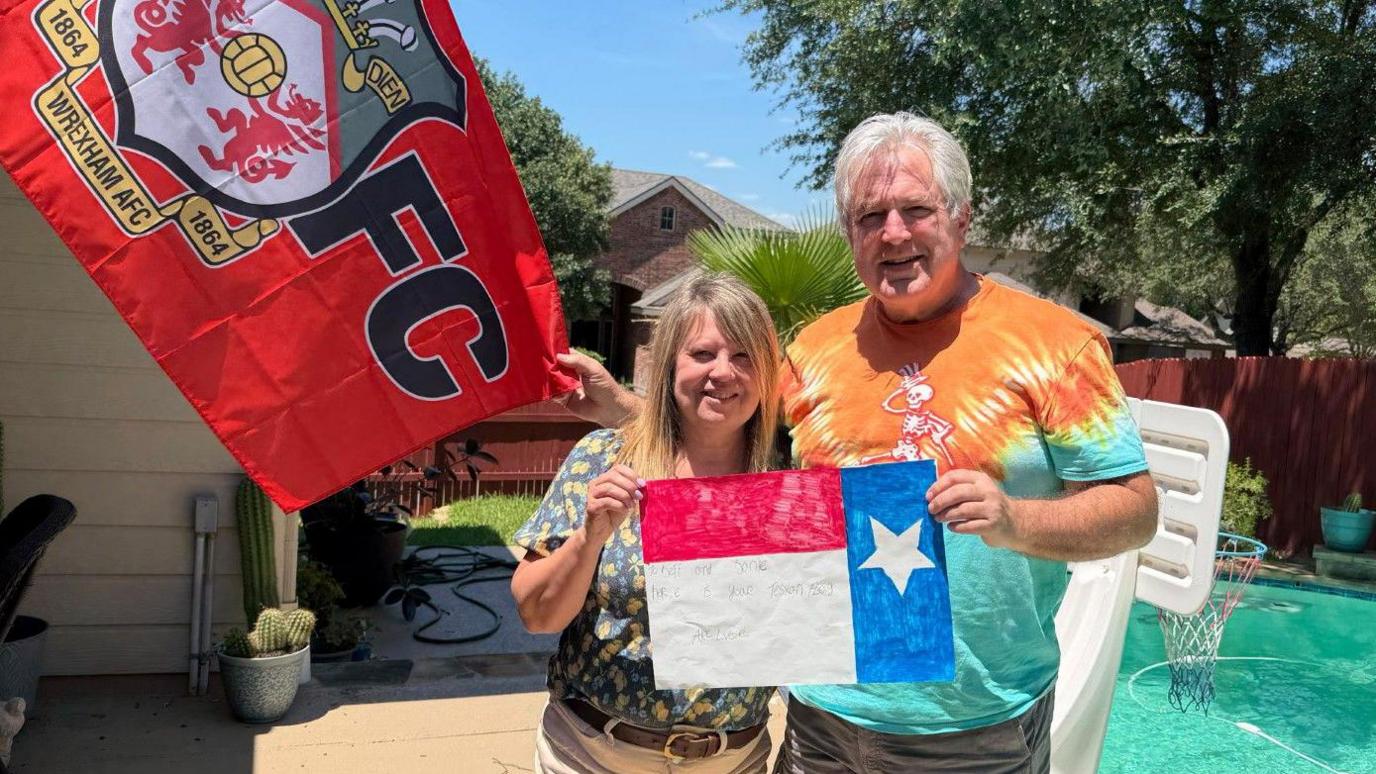A man and a woman in their early 60s, wearing shorts and t-shirts, stand in the sunshine in front of a small back garden swimming pool. They are holding a painting of the Texas flag between them and there is a  large Wrexham AFC flag behind them to the left. The woman has blonde shoulder-length hair and the man has short white hair. They are both smiling. Trees, plants, a fence and a neighbouring house are visible in the background.