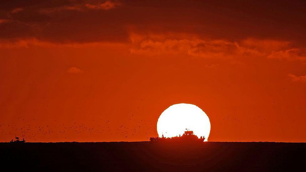 The Sun sets over an ocean. The sky is dark red and the silhouette of a ship sailing across the ocean in front of the Sun.