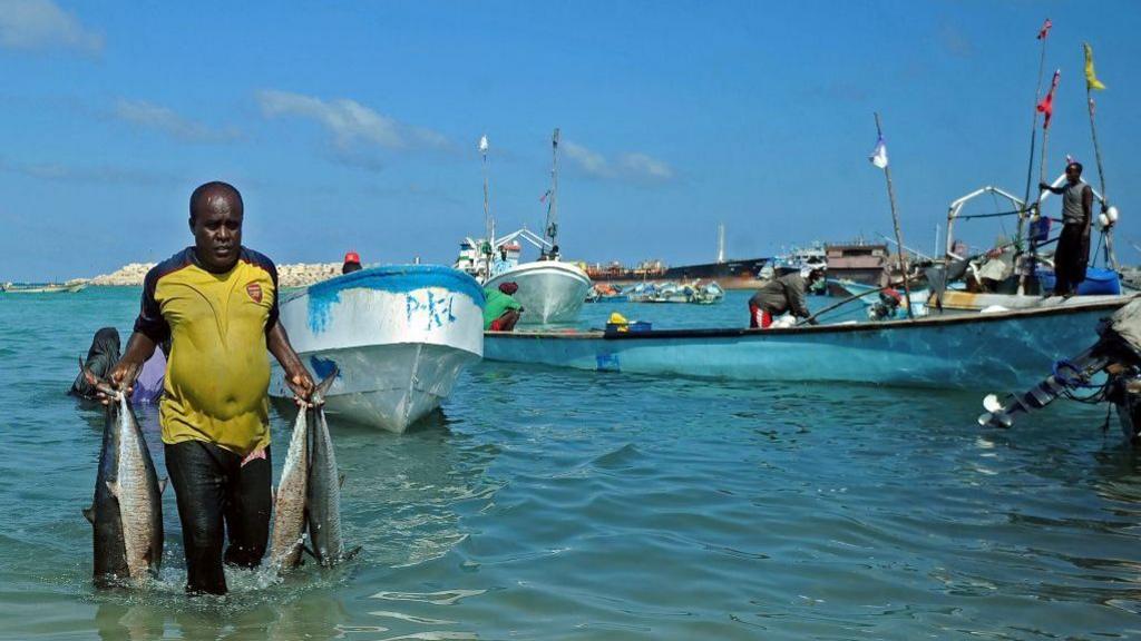 A man in a yellow top in the left of the picture wades through water holding four large fish in his hands. Fishing boats and the ocean can be seen behind him.