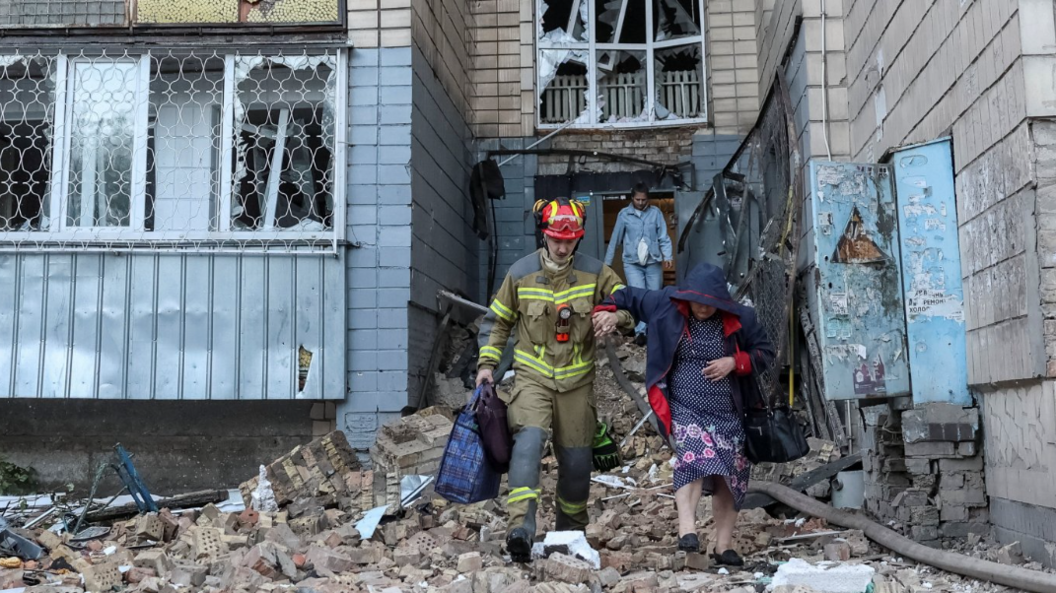 A firefighter helps an old lady over rubble in front of a destroyed building