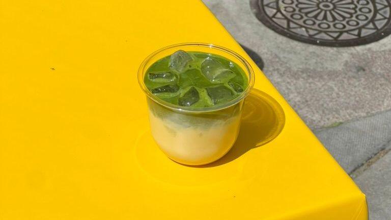 A plastic cup, half filled with a yellowy cream and half filled with bright green matcha and ice cubes, is pictured on a yellow table outside
