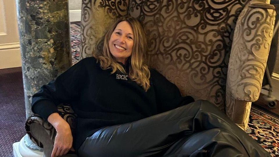 Vivienne Duval, a woman with dark blonde hair sits on an armchair with her legs crossed. She is smiling at the camera