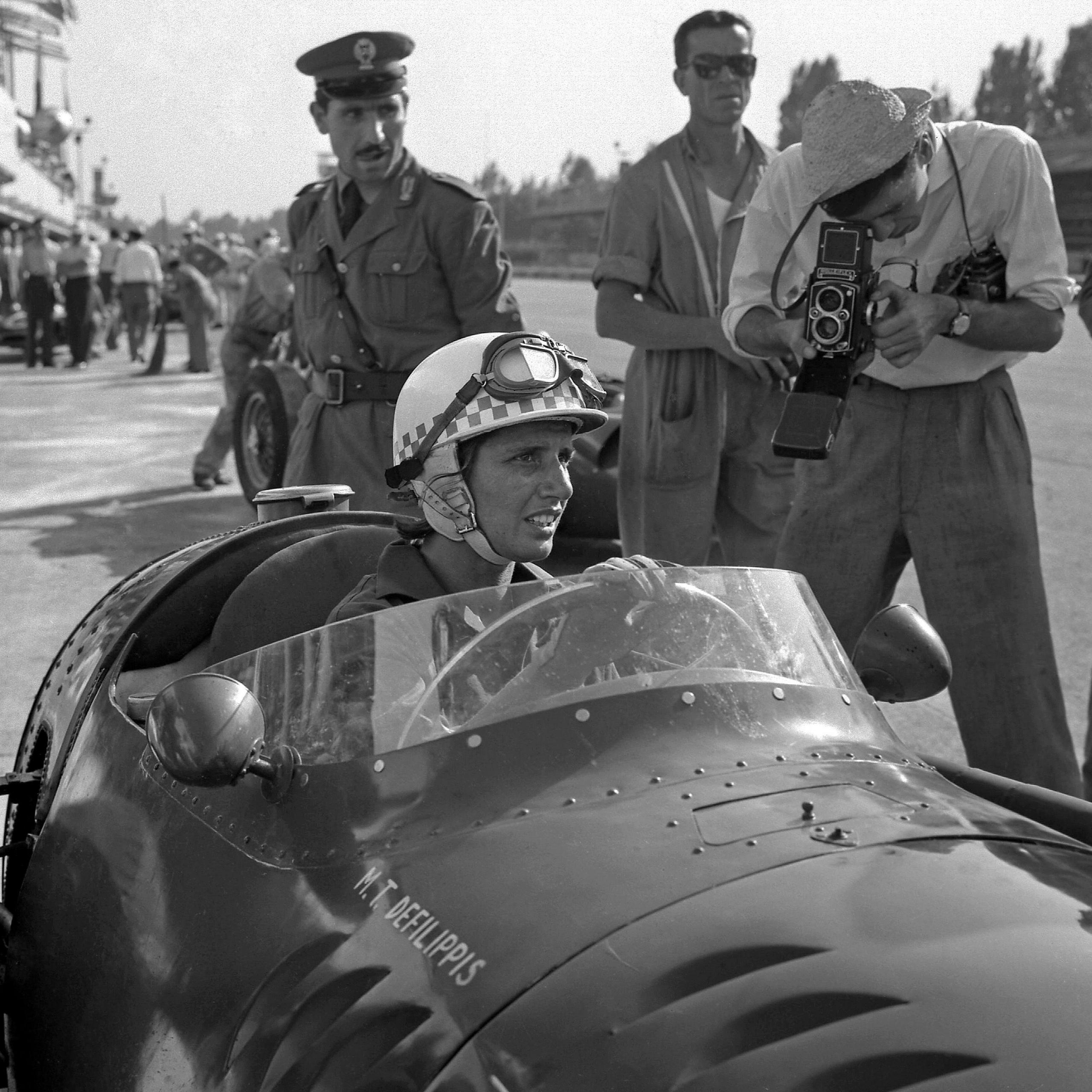 Maria Teresa de Filippis - the first woman at compete in F1 - sat in her Masreati while a photographer is stood behind her taking a picture
