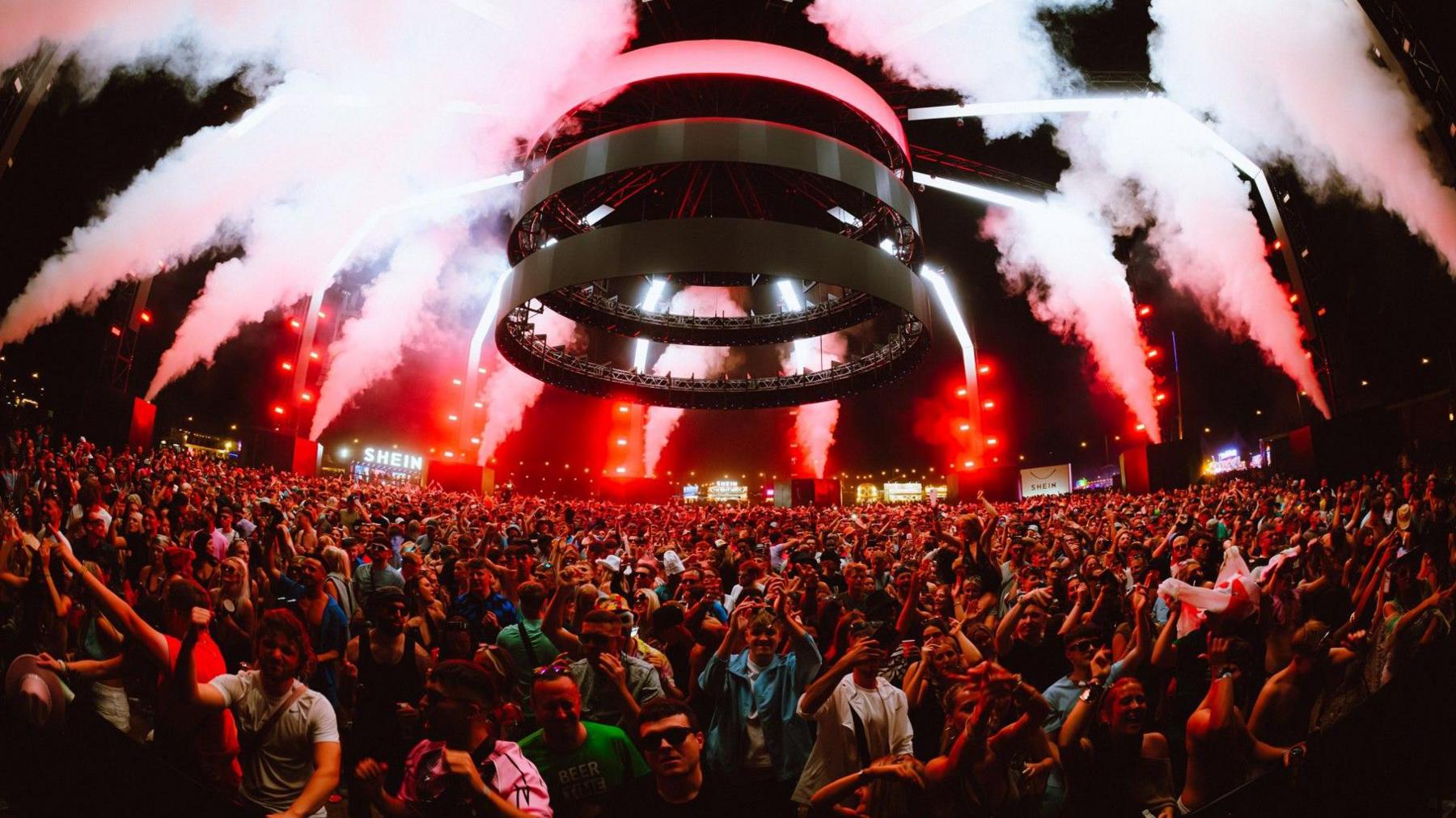 A large circular structure above a large crowd of revellers. Red lights and smoke can be seen from the edges of the venue.