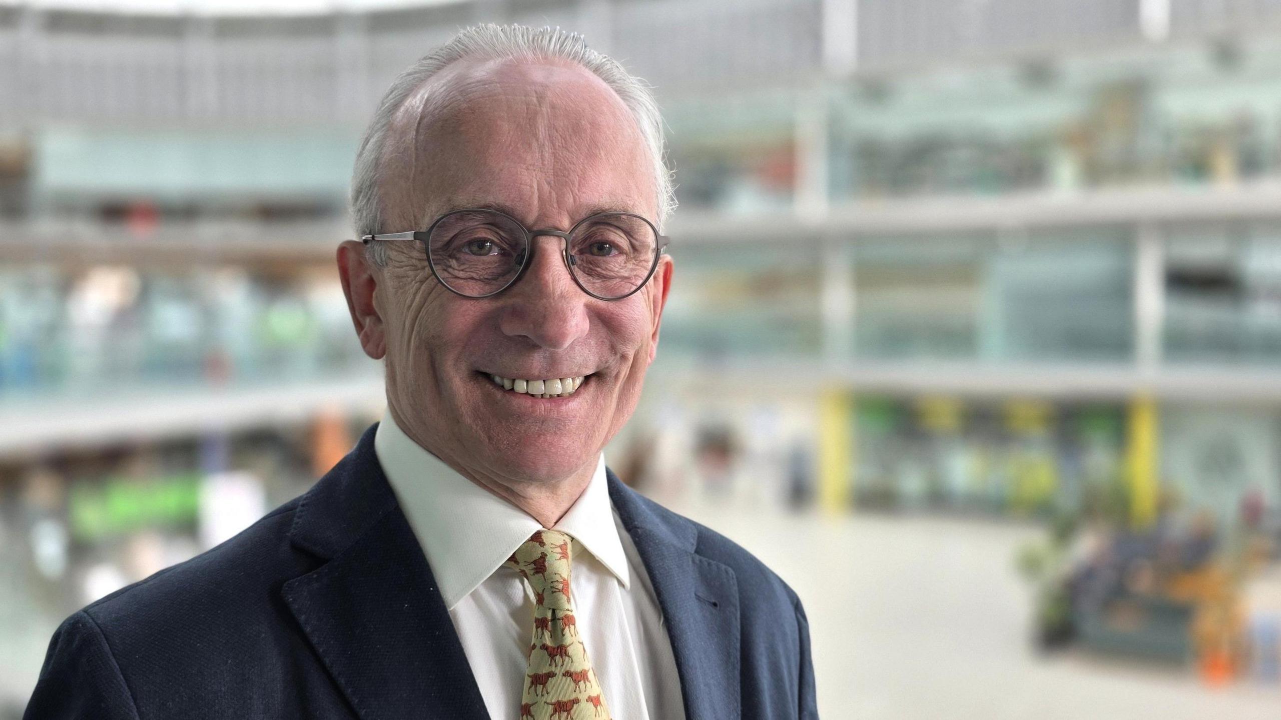 David Bick is smiling. He is wearing a navy suit jacket over a white shirt and patterned tie. The inside of the Norwich Forum is behind him but out of focus.