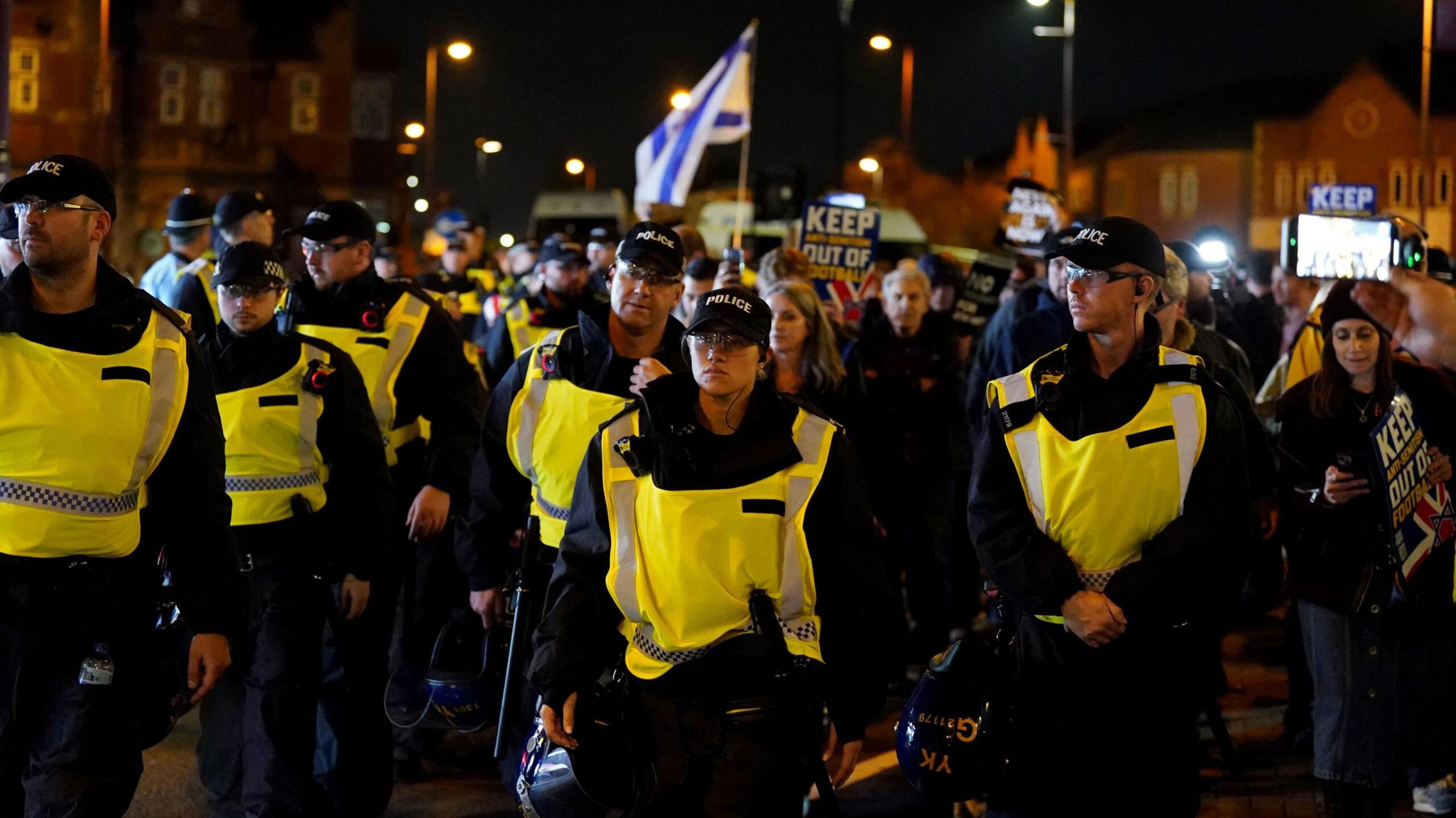 A group of officers are walking down a street outside Villa Park, most are male but in the middle is a white female officer wearing a black cap, all of them are wearing hi-vis vests