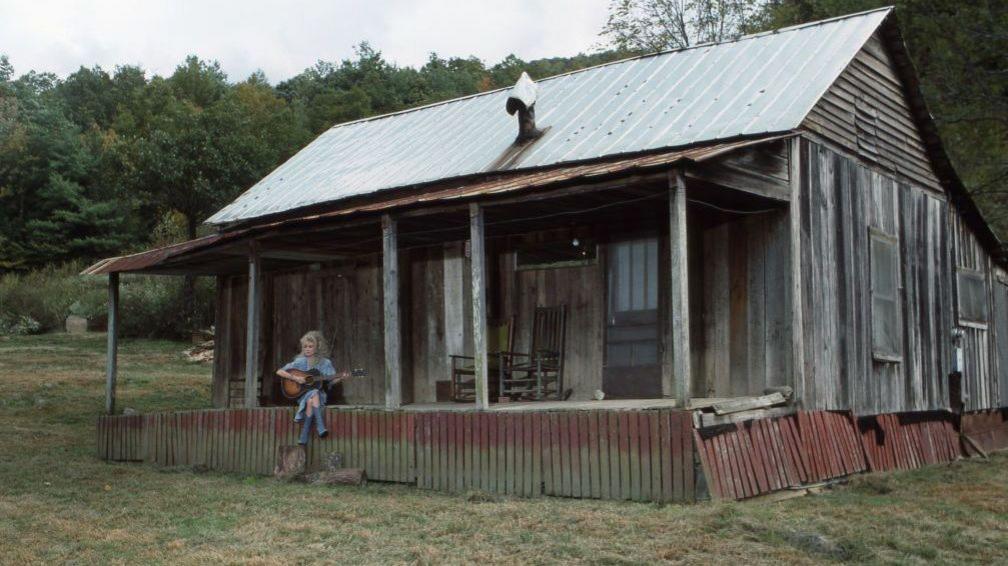 Dolly Parton sits on the porch of a replica of the log cabin she grew up with