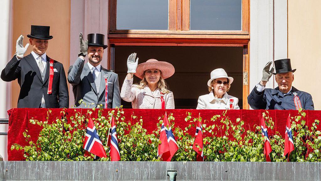 Three men and two women dressed in hats and formal wear stand on a balcony waving, flags in front of them.