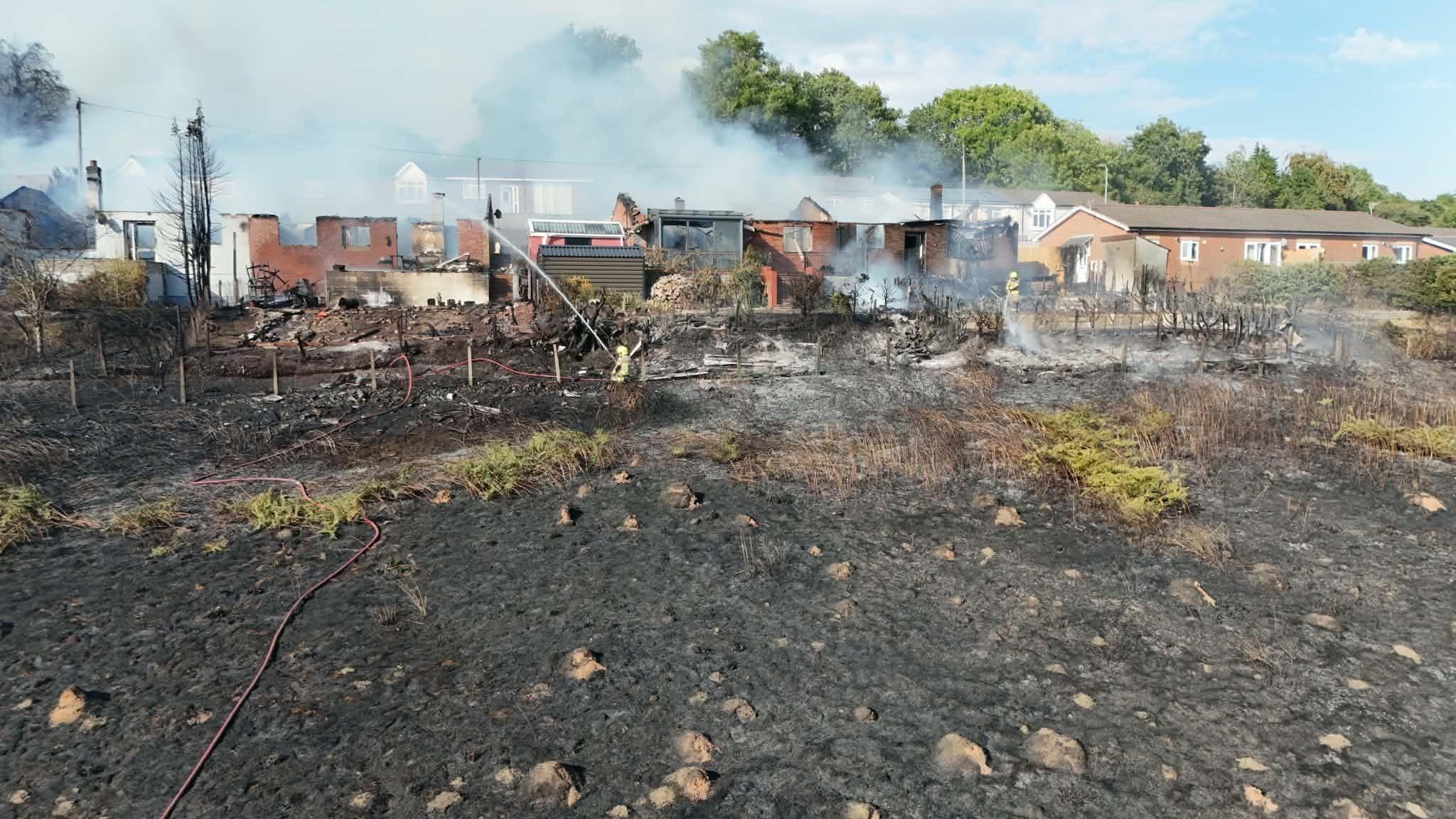 burned out grass in the foreground with smoke billowing from roofless black bungalows damaged in the fire