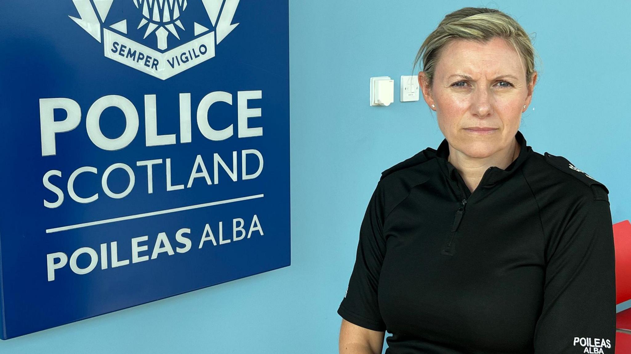 Ch Supt Emma Croft wears a black police uniform and sits in front of a light blue wall next to the dark blue Police Scotland crest