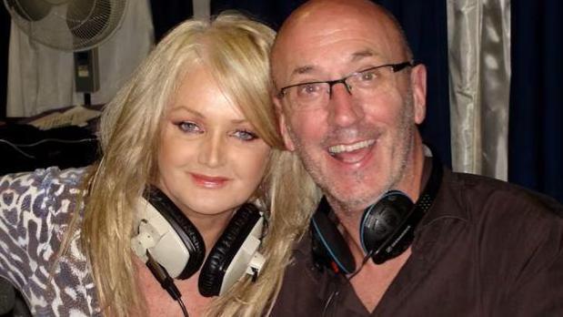A woman with long blonde hair sat down next to a bald man wearing glasses. They both have headphones around their necks and smiling at the camera.