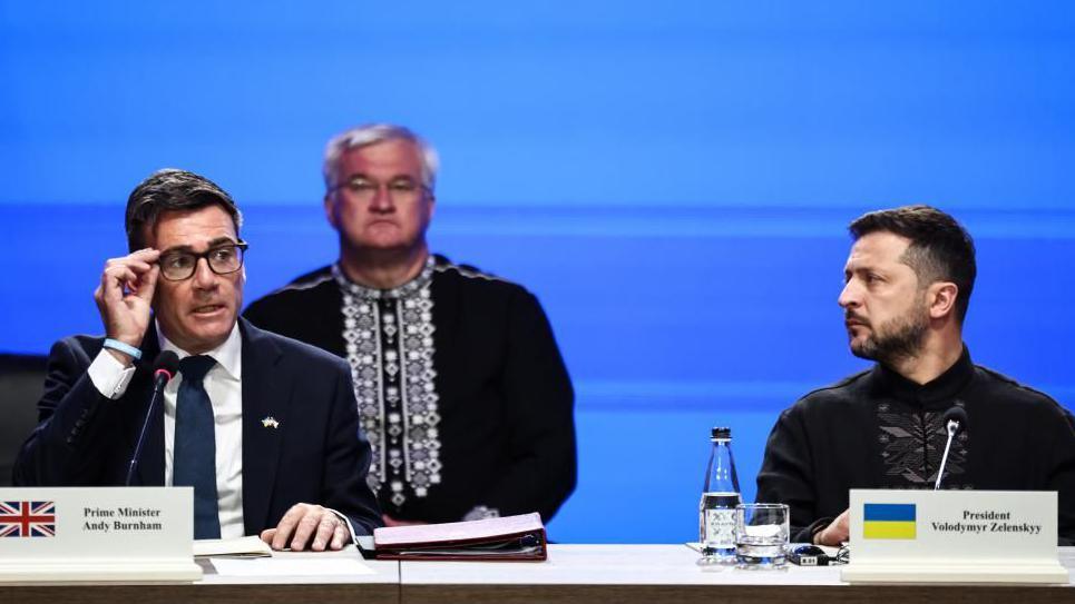 Andy Burnham adjusts his spectacles on the left of the image as Ukrainian president Volodymyr Zelensky looks across at him. The pair are on a stage and have name plates and folders in front of them.