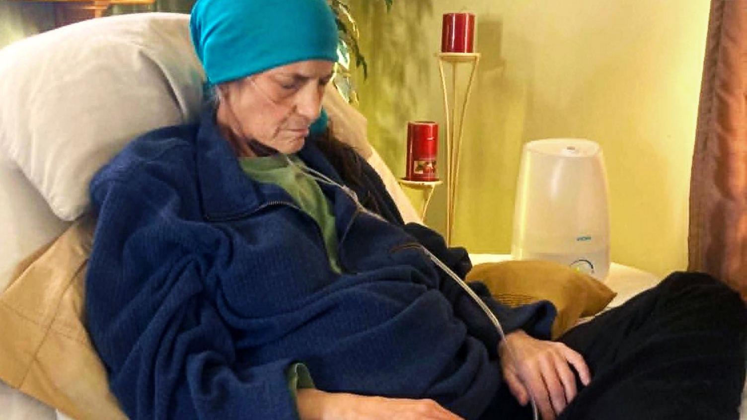 Kelly lies on a hospital bed propped up by pillows and looking down, as if asleep. She wears a blue headscarf on and has a clear tube feeding into her nose. Her hands rest of her lap.