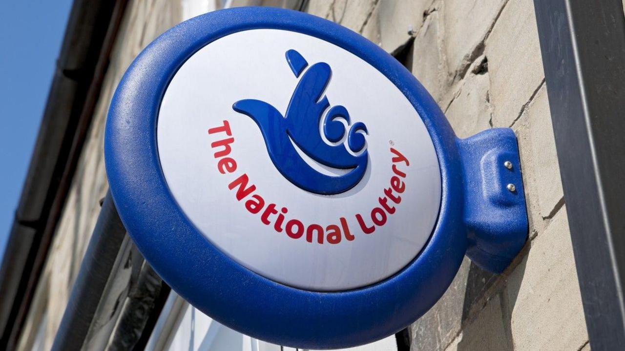 A shot of a sign attached to a wall with "The National Lottery" printed on it. Above the words is an image of a blue hand with its fingers crossed.