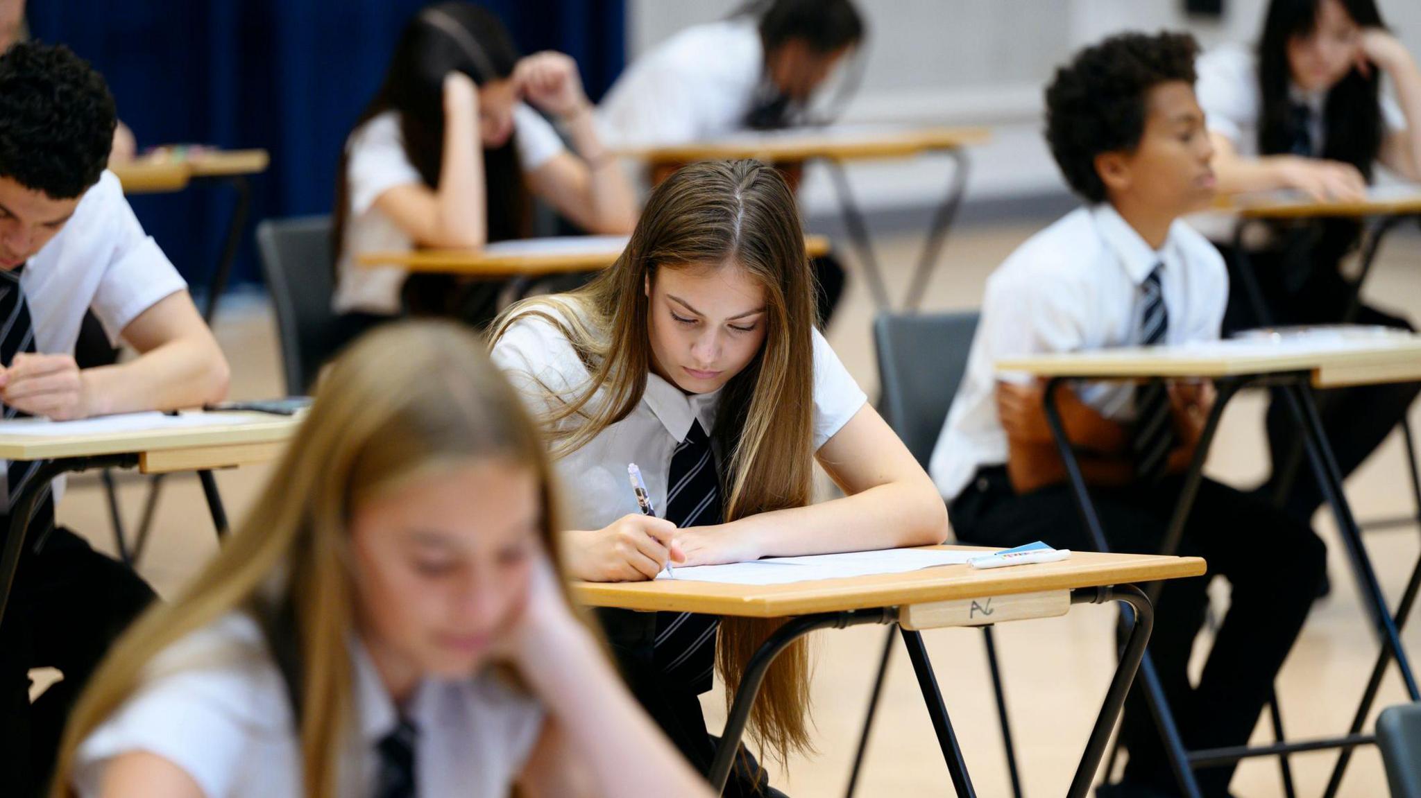 Teenage girl wearing school shirt and tie writing in exam booklet, arm resting on desk