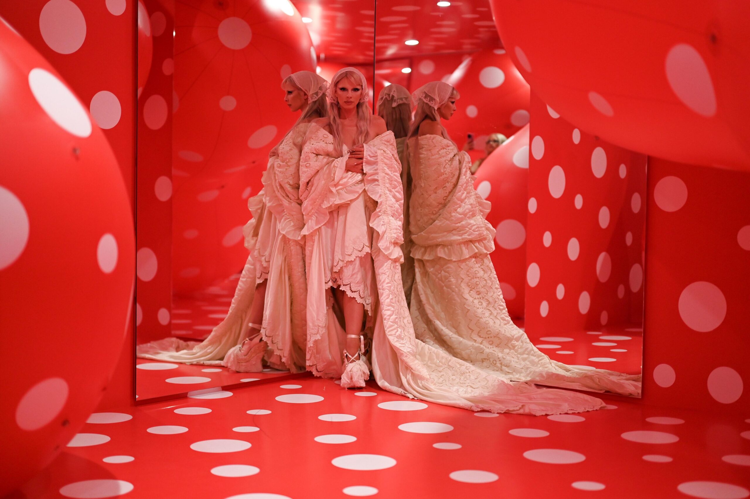 Julia Fox in an elaborate white dress looking at the camera and reflected in mirrors in a room with red walls and giant round sculptures covered in white spots