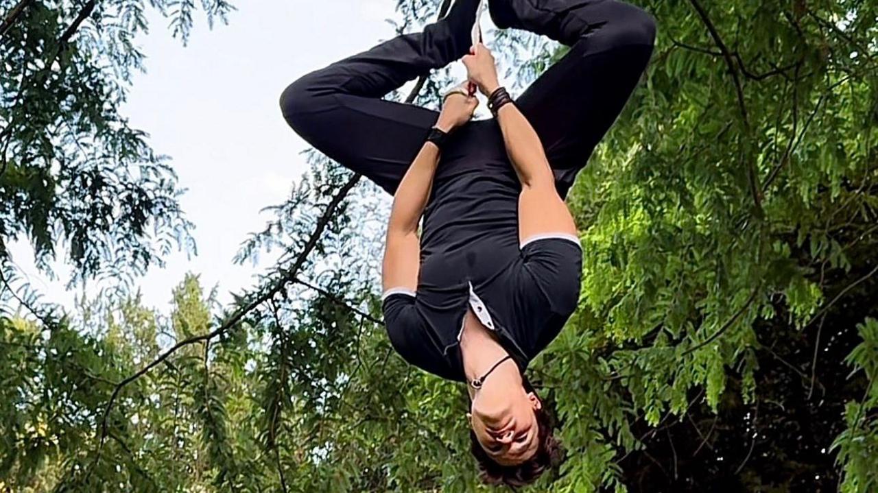 A man hangs upside from a rope with his legs apart at the knees. He's looking down. Behind him are trees and a blue sky.
