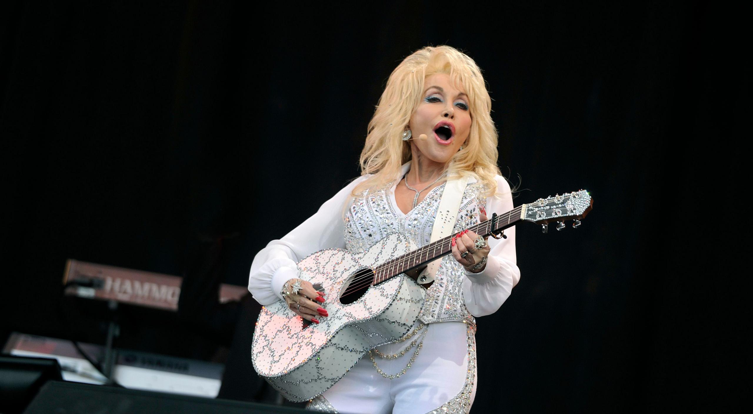 Dolly Parton, wearing a bedazzled white pants suit, strums a white guitar with pink fingernails, her mouth wide open in song under her flowing blonde locks