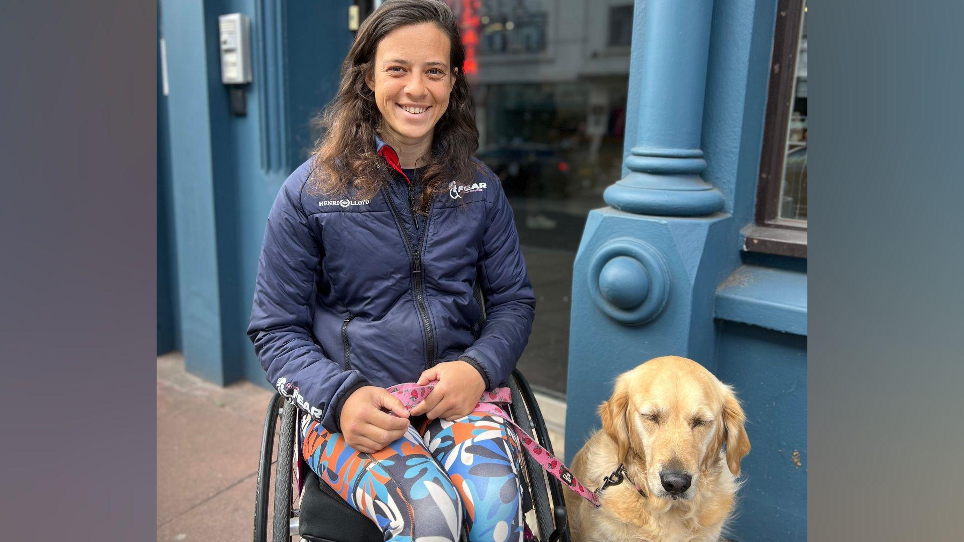 A woman wearing a blue puffer jacket and jogging trousers. She is sitting in a wheelchair and there is a golden retriever next to her.