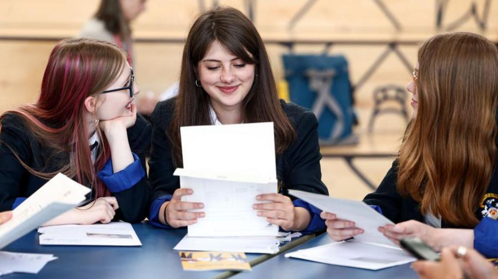 The photo shows three schoolgirls seated at a table looking at their exam results. They are wearing dark blazers with white shirts and ties. The table is covered with papers, envelopes, and folders. In the background, there are stacked chairs and a light-coloured wall, giving the impression of a hall or multipurpose room.