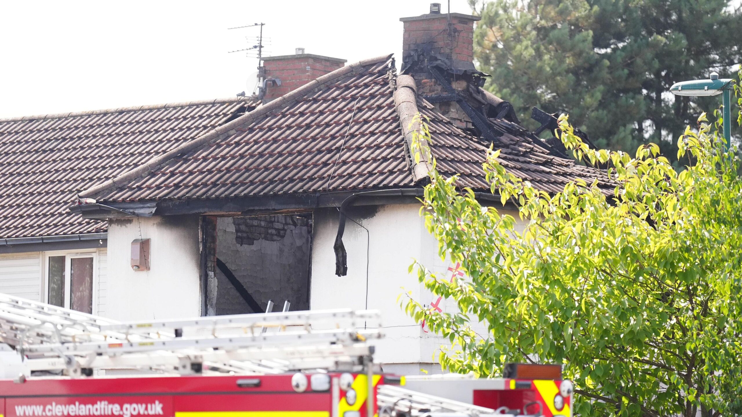 A fire engine is parked outside of the white house. The roof of the property is badly burnt, There are pieces of wood or metal sticking up out of it. The holes where the windows were are blackened.