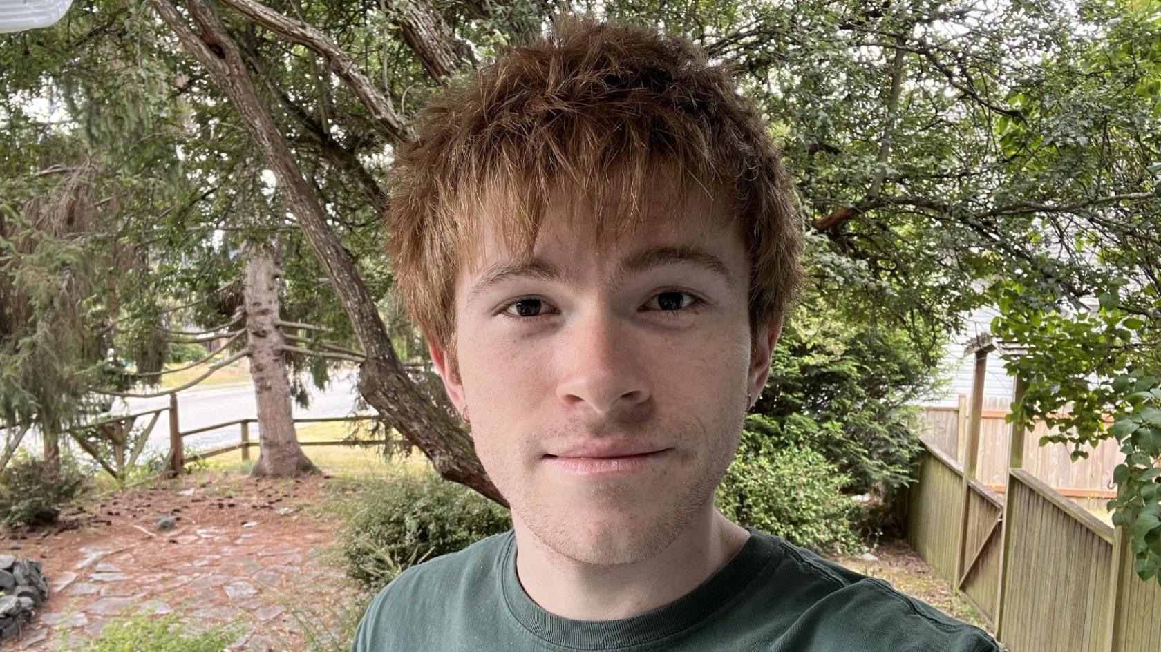 A man wearing a green T-shirt standing in the foreground. Trees, shrubs and a wooden fence fill the garden behind him. A stone path runs through the garden with a small pile of rocks visible near the path. A road and additional trees can be seen beyond the fence in the background.