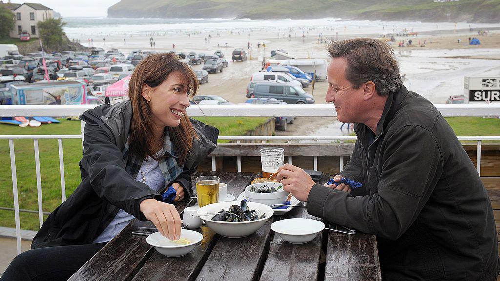 Samantha Cameron and David Cameron sitting opposite each other on a bench at a restaurant in Polzeath, Cornwall. They are drinking and eating mussels. They are smiling at each other. In the background is Polzeath beach and a car park.