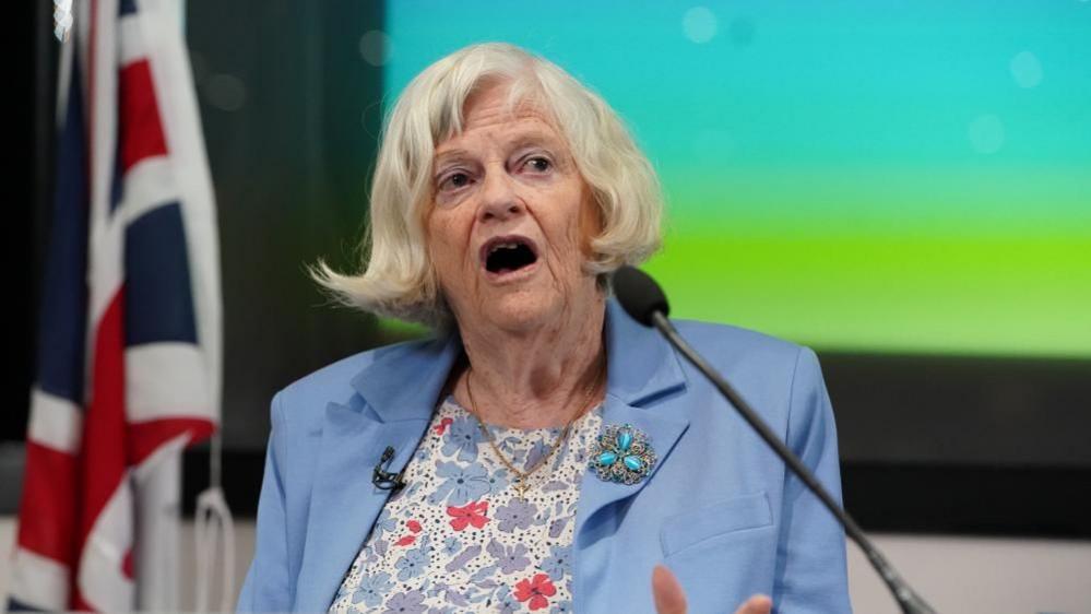 Ann Widdecombe is wearing a light blue blazer with a floral shirt and a turquoise brooch. Her hair is short and she is speaking in front of a microphone. The background behind her is blurred but a Union Jack can be seen.