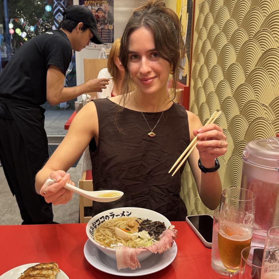 Faye Jawad, in a brown top, sits in a restaurant and poses with a bowl of ramen