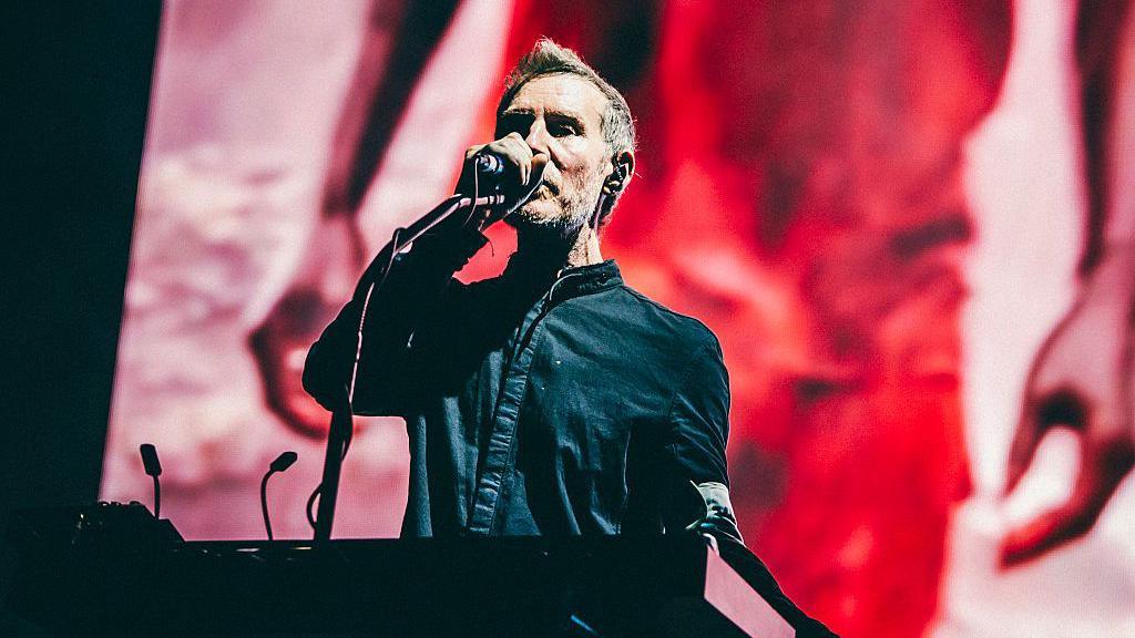 Robert Del Naja, one of the band members of Massive Attack, sings into a microphone while standing on stage at a keyboard