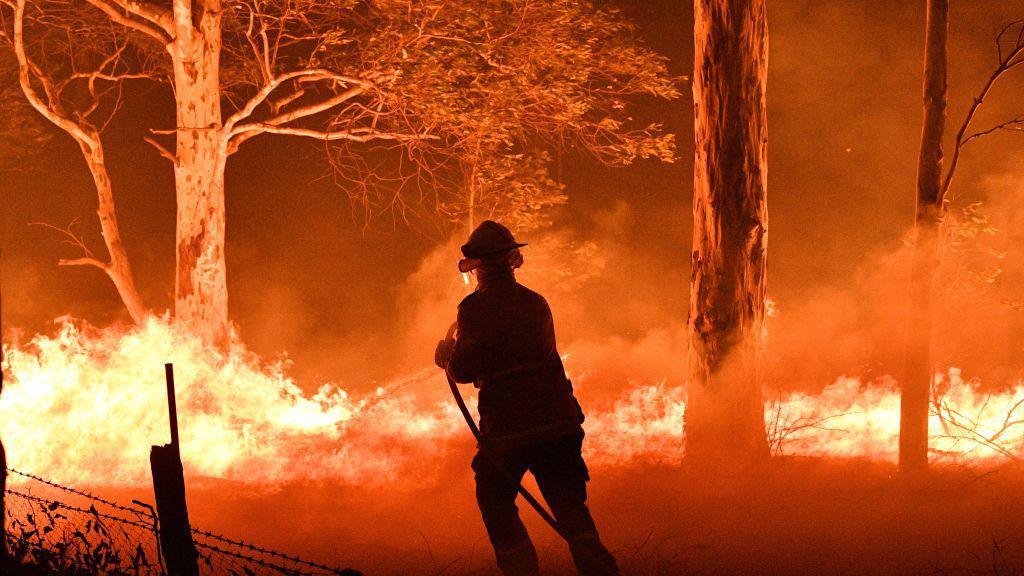 A firefighter using a hose on a major bushfire with bright flames covering the ground