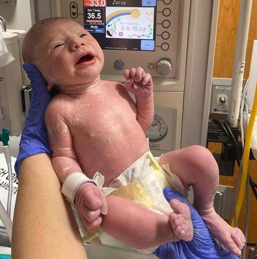 Newborn baby in the foreground, wearing only a disposable nappy and supported by gloved hands in front of a neonatal warming unit. A hospital identification band is attached to the baby’s wrist. The warming unit’s display screen shows temperature settings and a weight reading, with control buttons and gauges visible around it. Medical equipment, tubing and wall-mounted hospital fittings appear in the background. The image is taken in a clinical hospital setting.