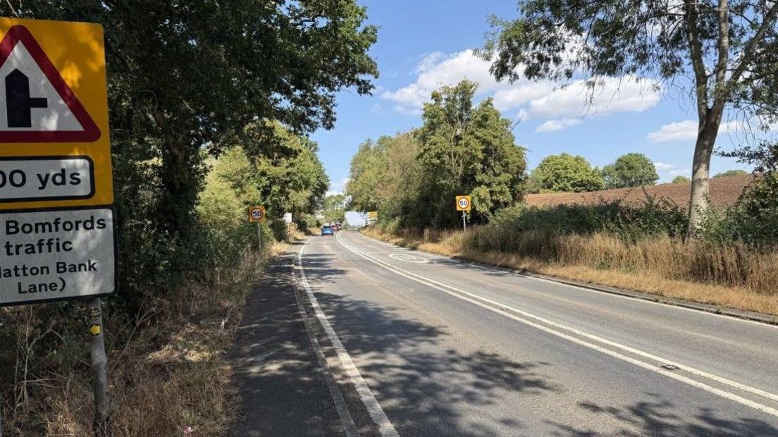 A stretch of road in the sunshine. A 50mph sign can be seen. The road is surrounded by trees with fields to the right hand side.