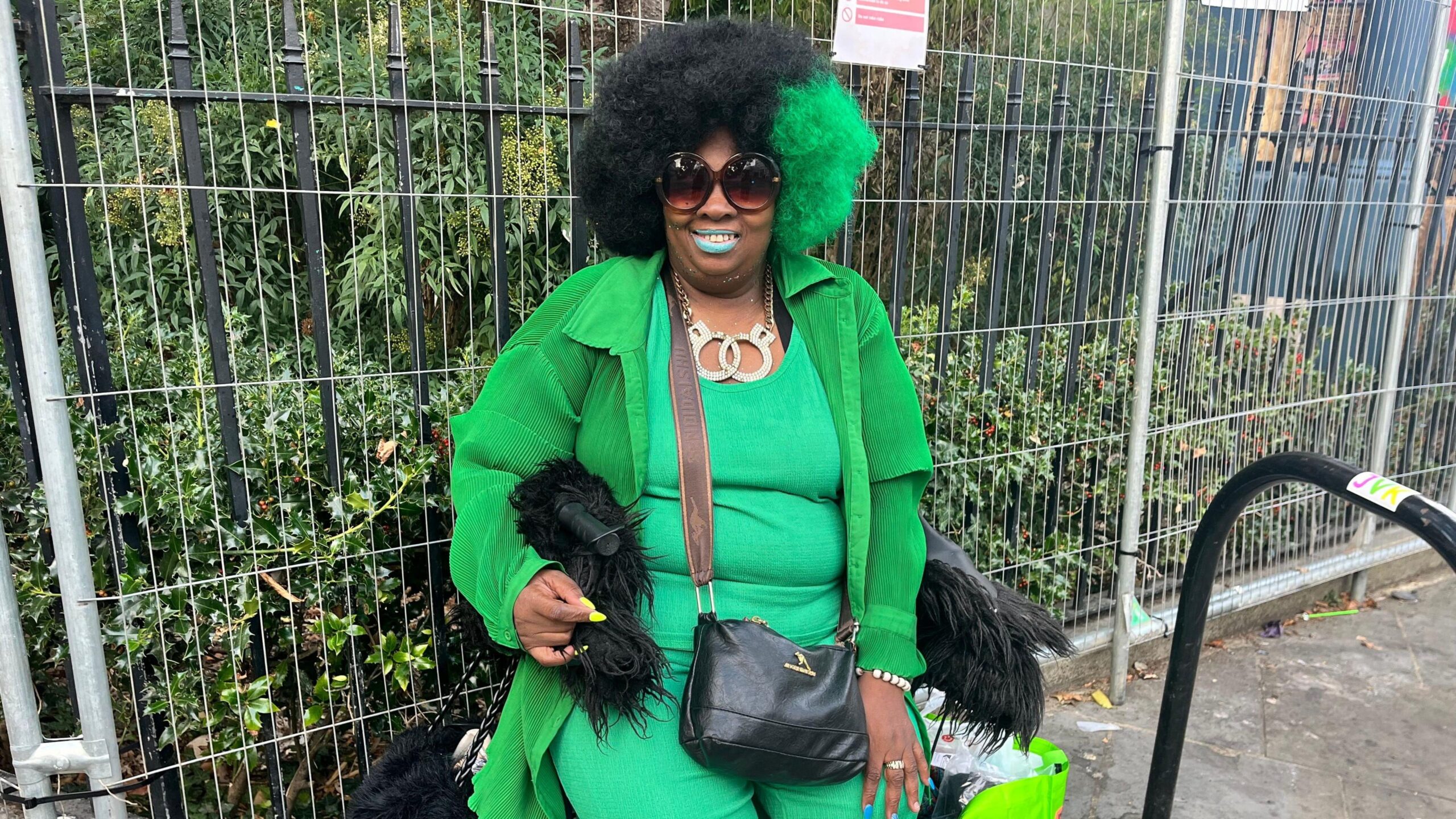 A woman in a green outfit with black sunglasses and a black bag sits on a chair on the pavement.