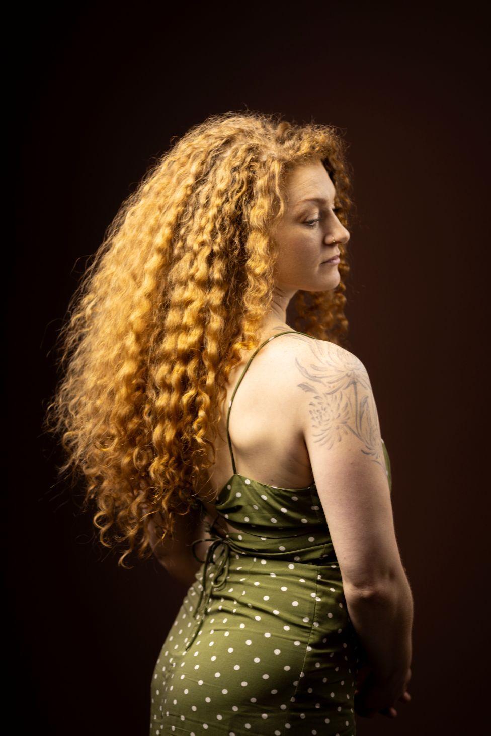 Studio portrait of a person with long, voluminous curly hair photographed from the back and side, turning slightly toward the camera. The person wears a green sleeveless dress with white polka dots and thin shoulder straps, featuring a lace-up back detail. A floral tattoo is visible on the upper arm. Warm lighting highlights the texture of the hair against a dark brown background.