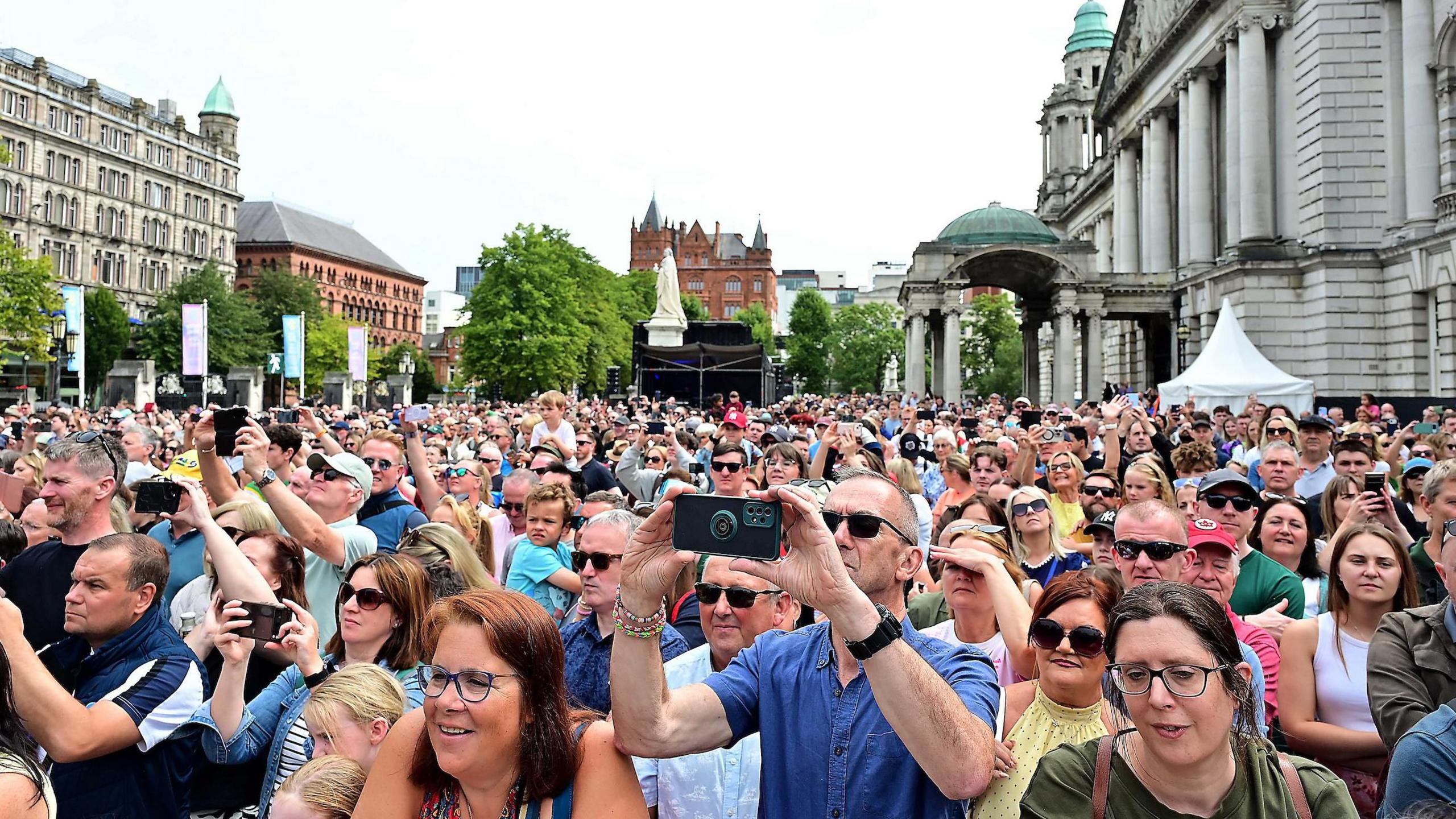 A large crowd of people stand in the grounds of Belfast City Hall, which is surrounded by tall buildings.  Many are wearing sunglasses and are using mobile phones to record what they see on a stage in front of them.