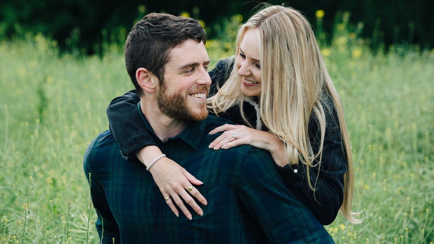 Andrew and Lissie Harper. She is standing in front of her looking back as she wraps her arms round his shoulders. Both are smiling