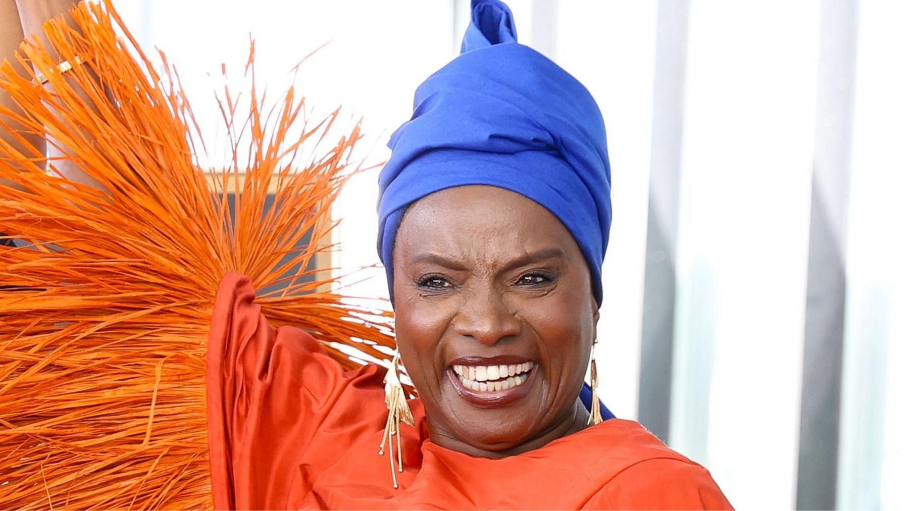 Angélique Kidjo smiles and lifts one arm in a long-sleeved dress with tasselled sleeves and a royal blue headwrap